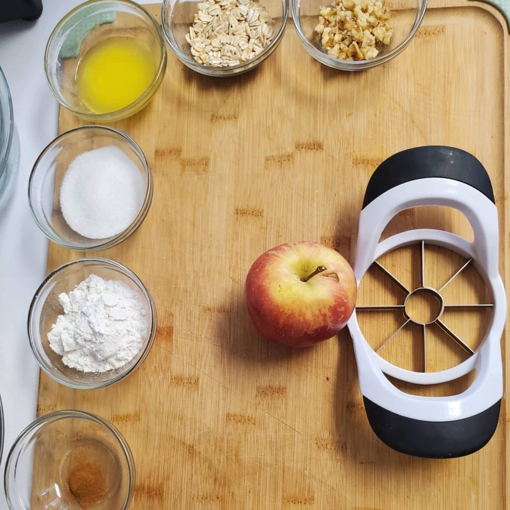 Cutting board with mise en place:  apple with apple corer, flour, sugar, cinnamon, butter, oats, walnuts