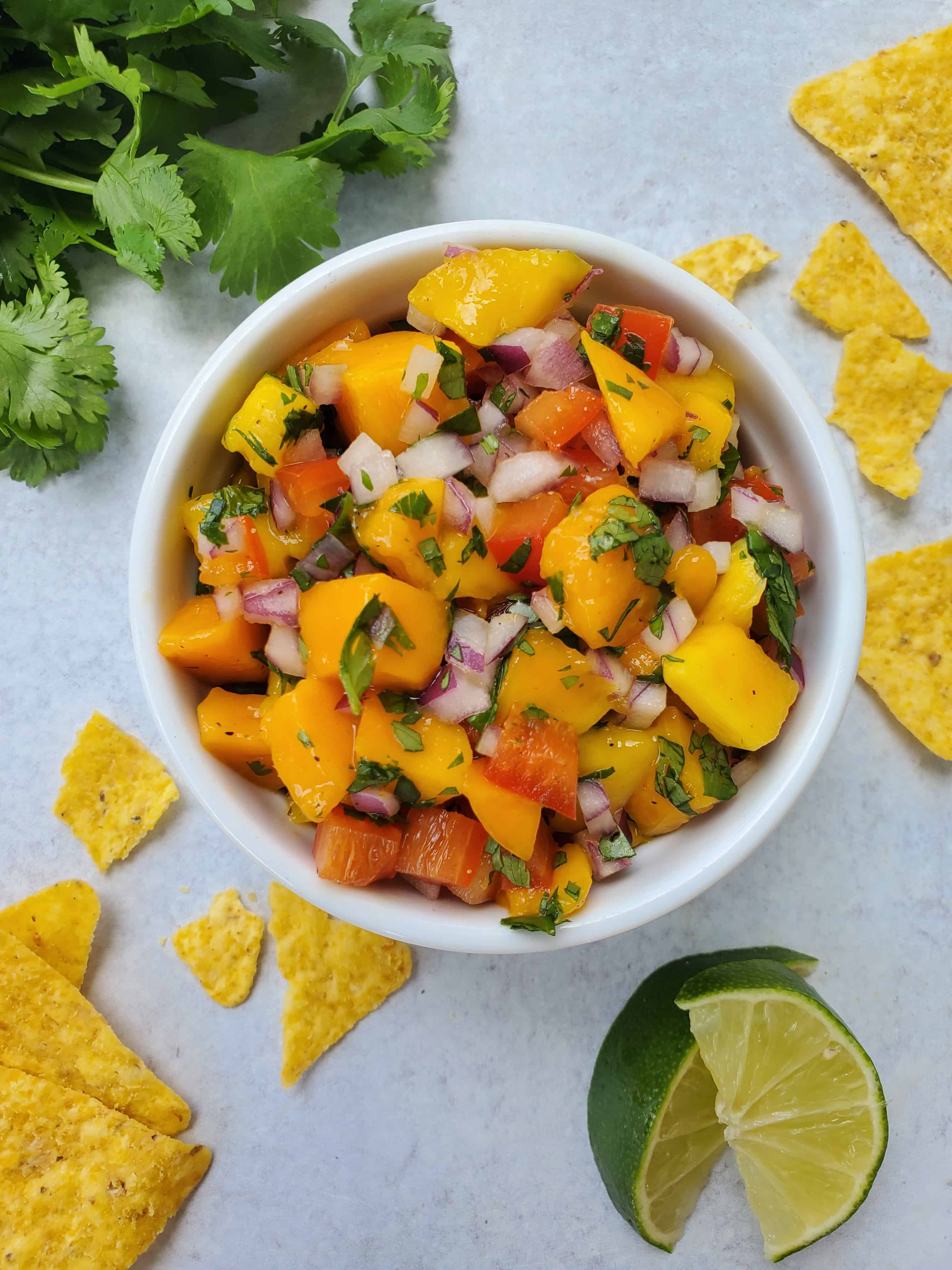 Mango salsa with mango, red pepper, red onion, cilantro in a bowl with chips, cilantro, and lime as garnish