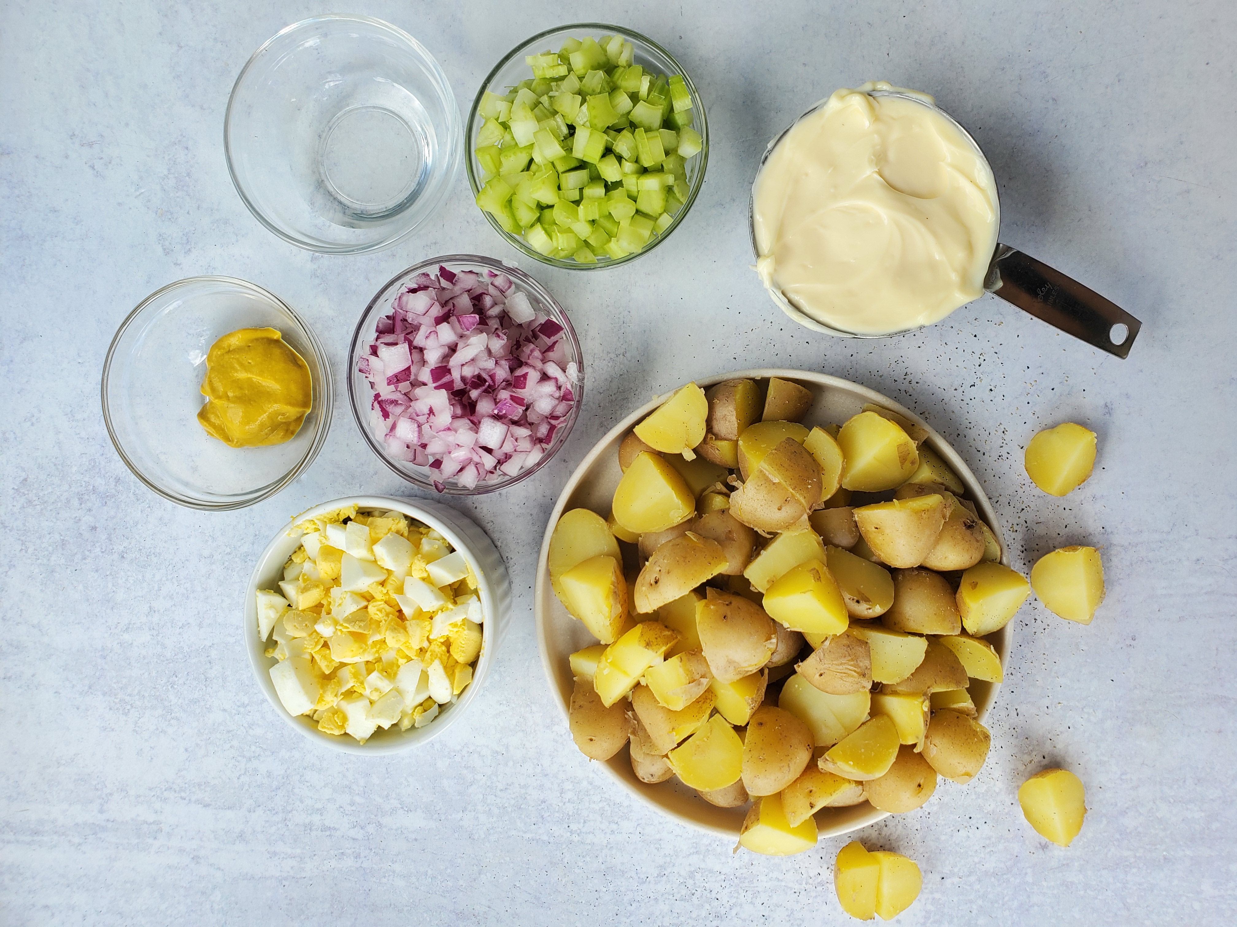 ingredients in a separate bowls for egg potato salad - potatoes, mayonnaise, celery, onion, hard-boiled egg, vinegar, mustard