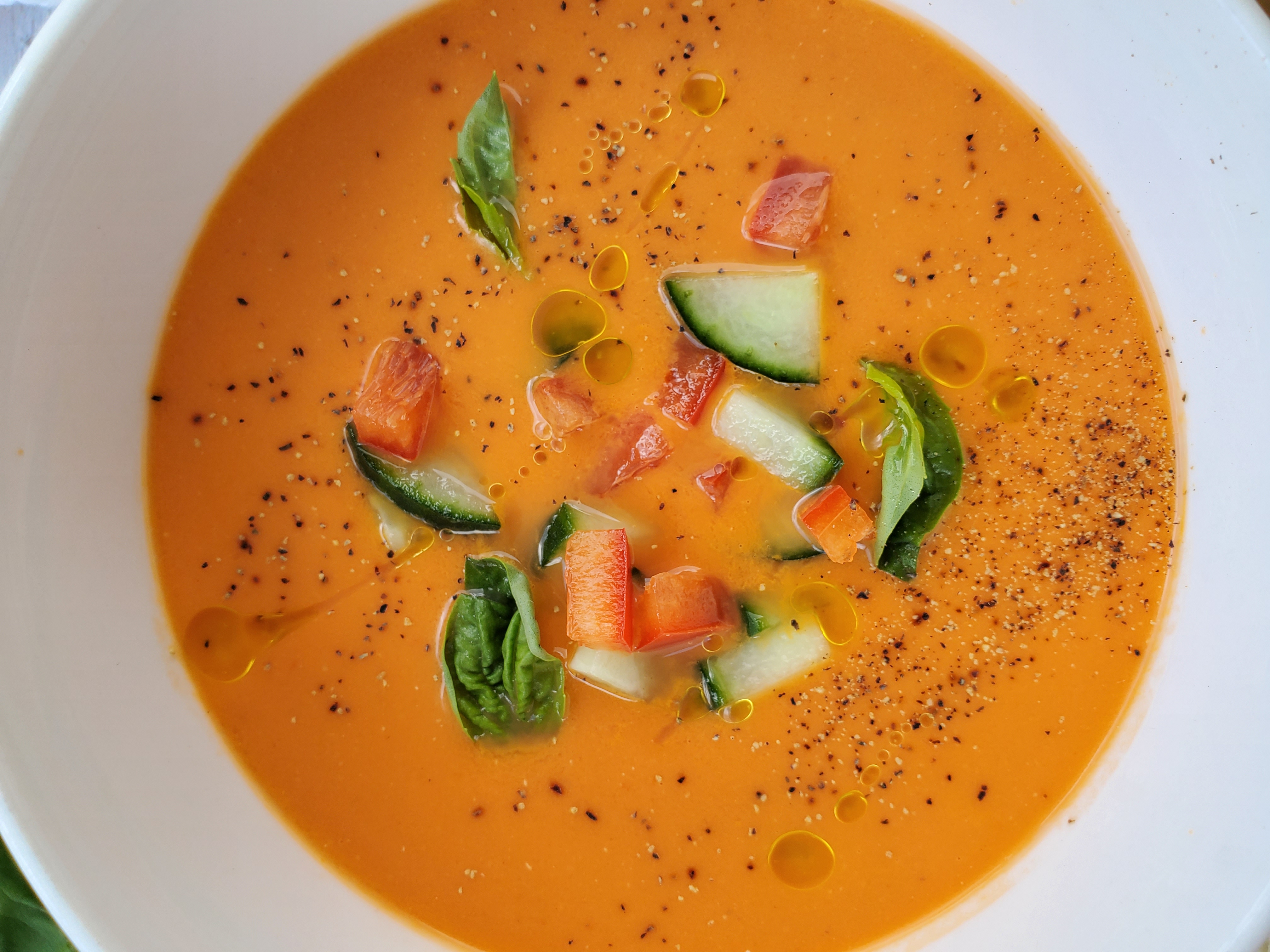 bowl of gazpacho up close and with a few pieces of cucumber, pepper, and basil sprinkled on top
