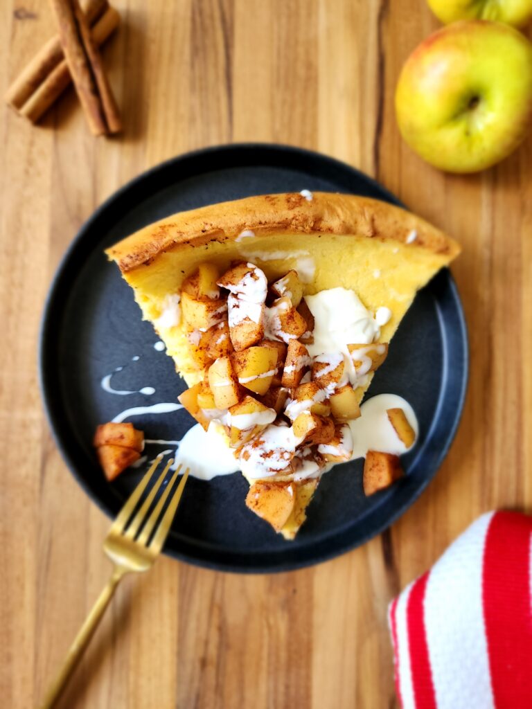 Single slice of a dutch baby on a plate topped with cinnamon apples and creme fraiche glaze