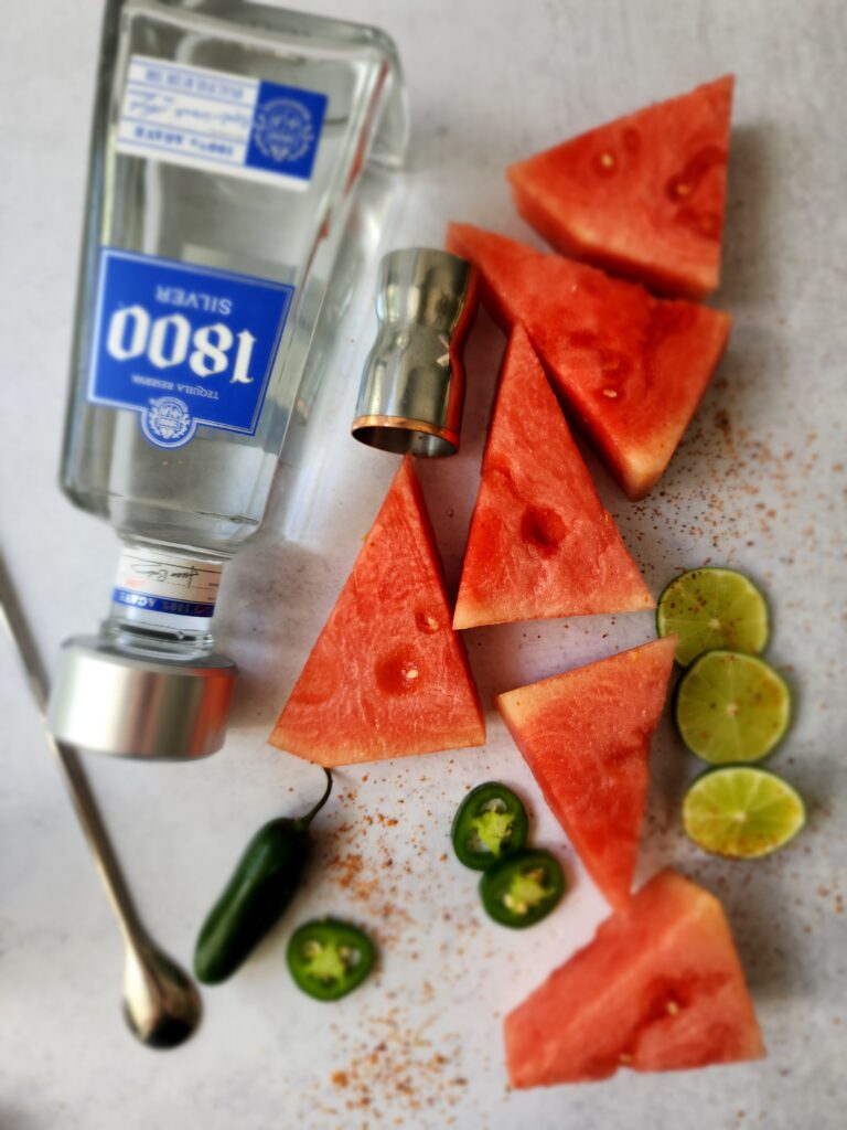 Top-shot of all the ingredients for a tajin watermelon margarita - tequila, simple syrup, jalapeno, watermelon, lime