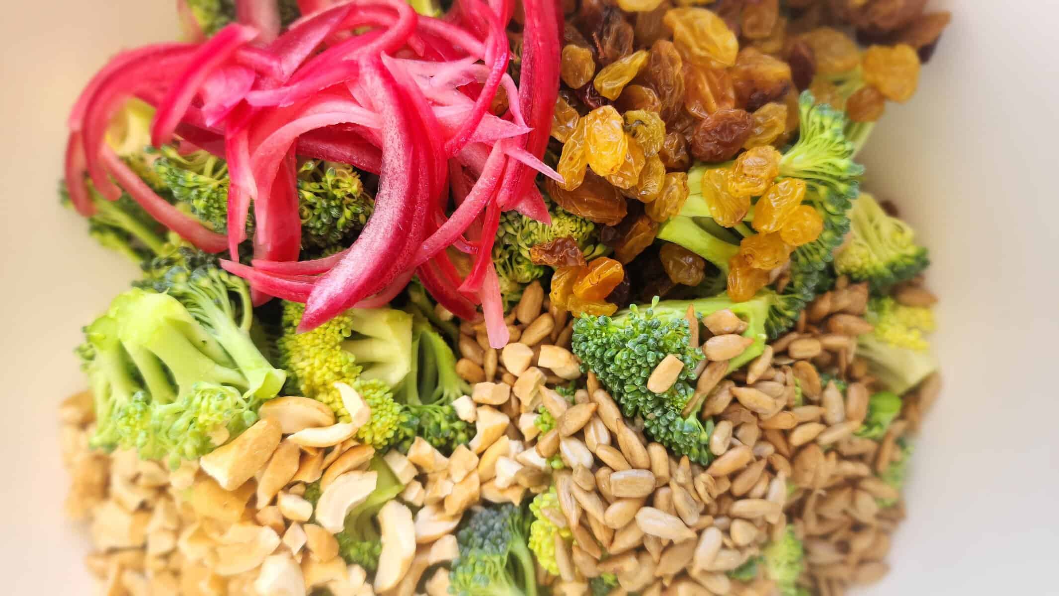bowl of broccoli salad ingredients:  broccoli florets, pickled red onion, sunflower seeds, cashews, and golden raisins
