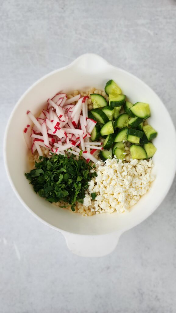 bowl of couscous with all the add-ins on top before being mixed: cucumber, radish, parsley, and feta cheese for lemon couscous salad