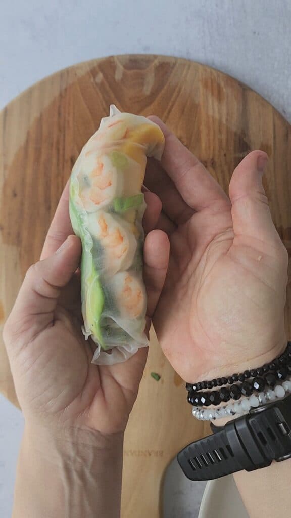 cutting board with my hands holding an easy shrimp spring roll close up with the shrimp showing through the wrap