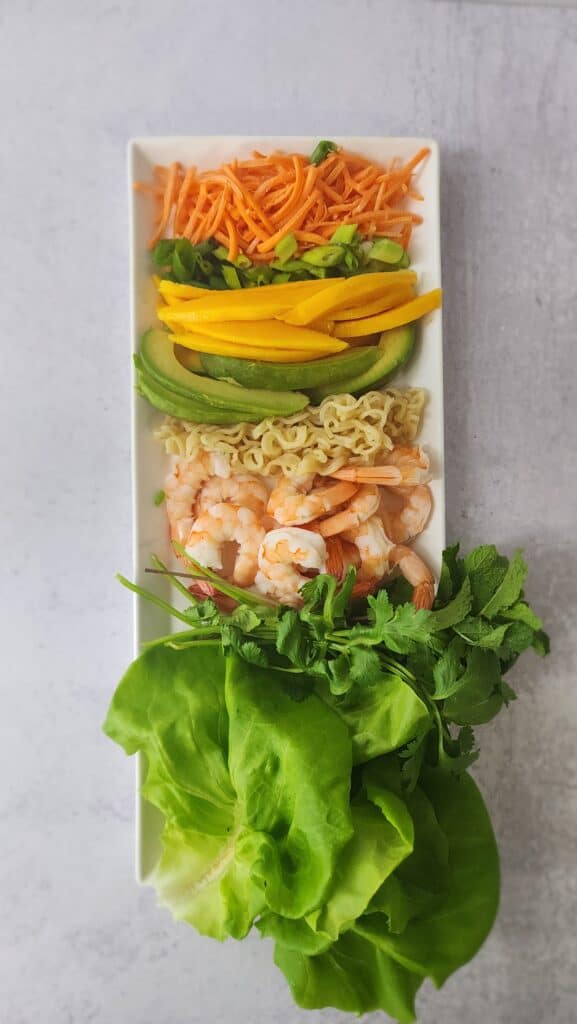 plate of ingredients for shrimp spring rolls: lettuce, herbs, cooked shrimp, avocado slices, mango slices, scallions, carrots