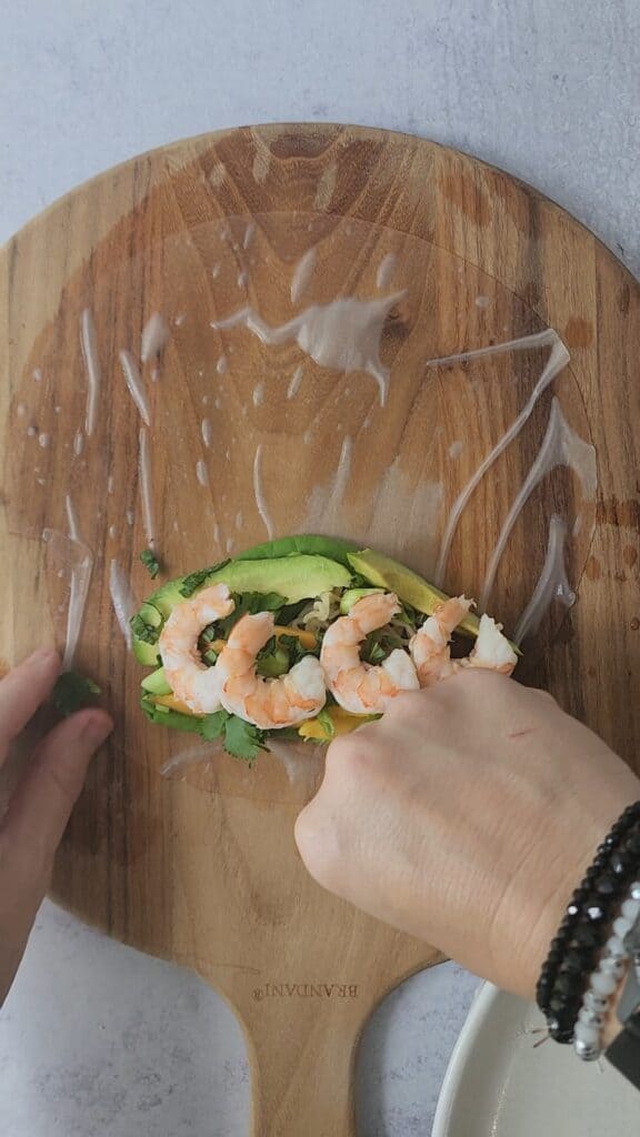 cutting board with a soaked rice paper with the layers for an easy shrimp spring roll: lettuce, noodles, avocado, shredded carrot, herbs, and cooked shrimp