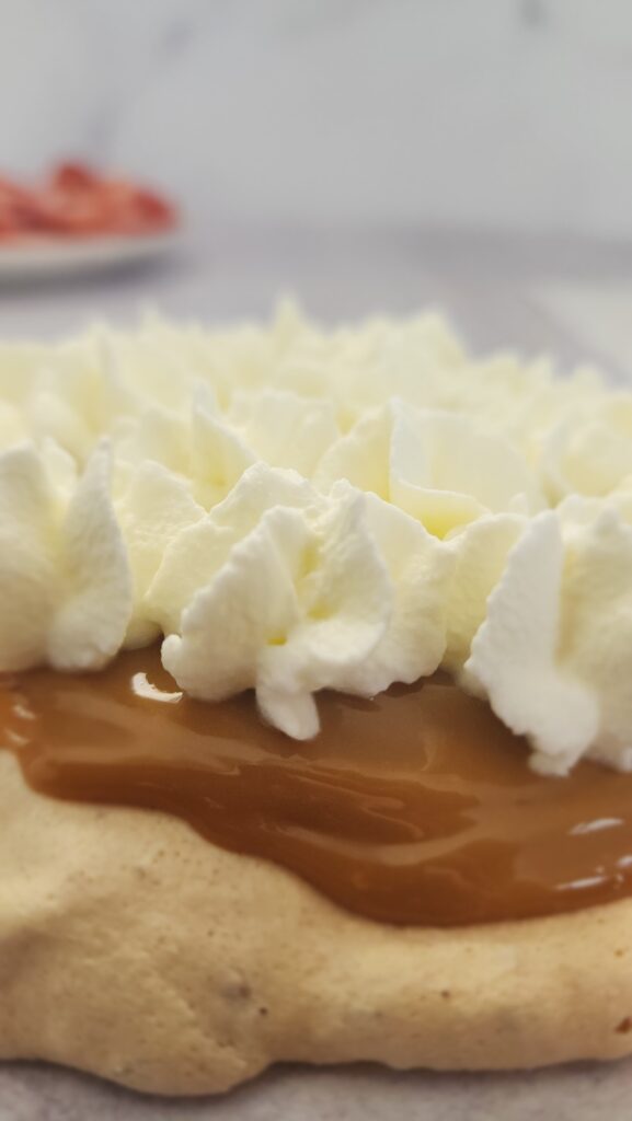 the makings of a strawberry pavlova layer by layer: here the pavlova has the dulce de leche spread on and now the whipped cream has been piped on