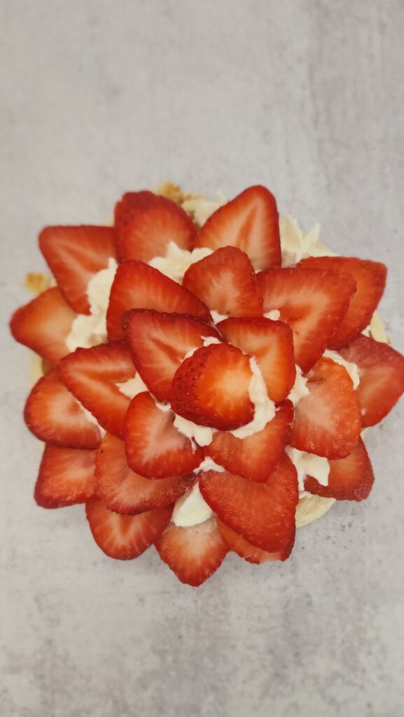 the makings of a strawberry pavlova and the pavlova is on the bottom with then the dulce de leche, next the whipped cream, and now the strawberries are being assembled