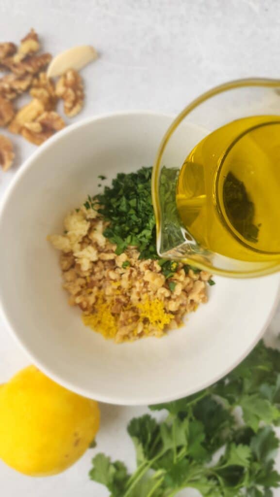 pouring olive oil into the bowl of parsley, walnuts, lemon zest and garlic for walnut gremolata