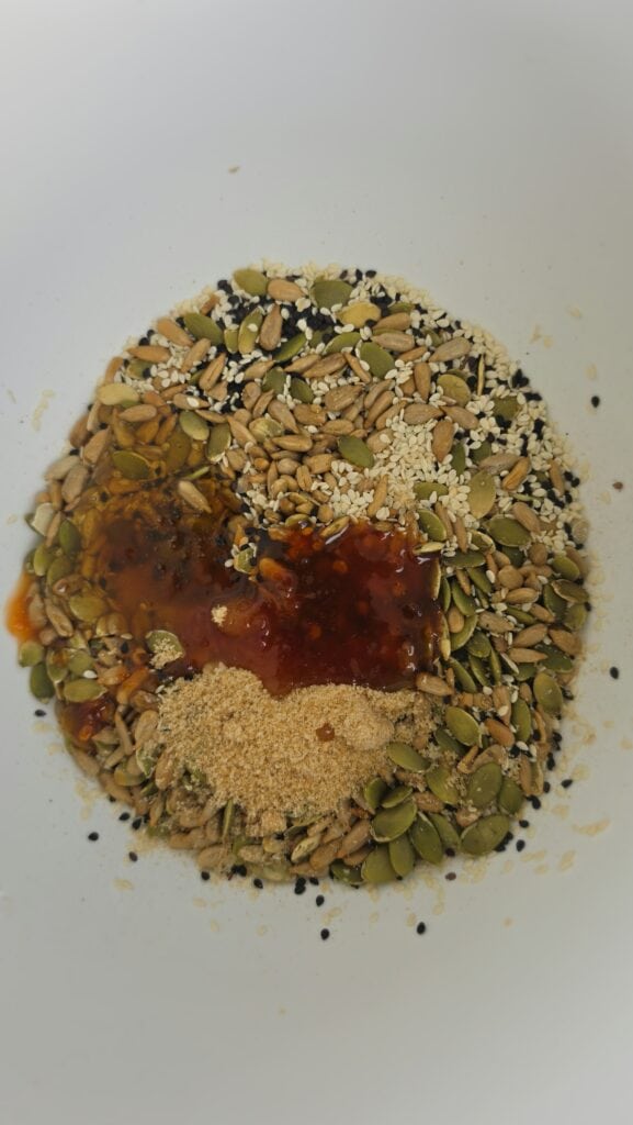 pumpkin seeds, sunflower seeds, white sesame seeds, and black sesame seeds in a bowl with chili sauce, brown sugar, honey, ready to be stirred together for seed brittle