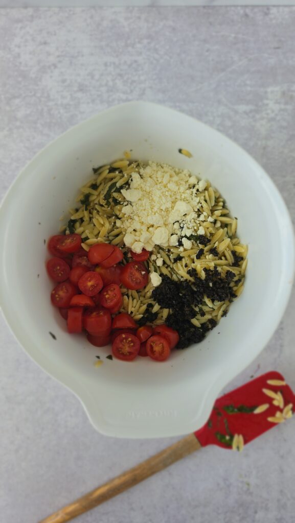 grape tomatoes, feta cheese, black olives in a bowl being added to orzo with wilted spinach for orzo salad